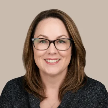 Woman with shoulder-length brown hair, wearing glasses and a dark blouse smiles in front of a neutral background.