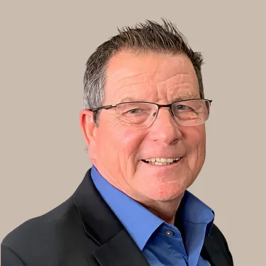 Man wearing glasses, blue shirt, and dark blazer smiles in front of a neutral background.