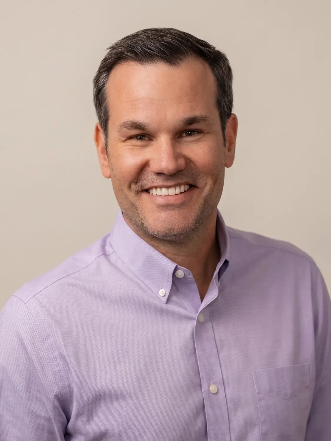 Smiling man with dark hair wearing a lavendar button-down shirt in front of a neutral background.