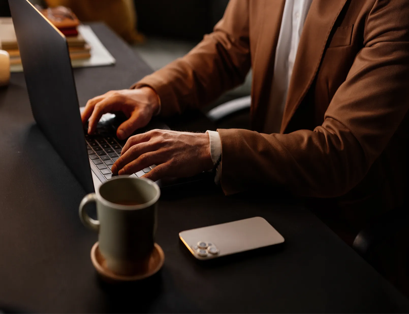 Person in a brown blazer typing on a laptop at a desk with a coffee cup and smartphone nearby, in moody lighting.