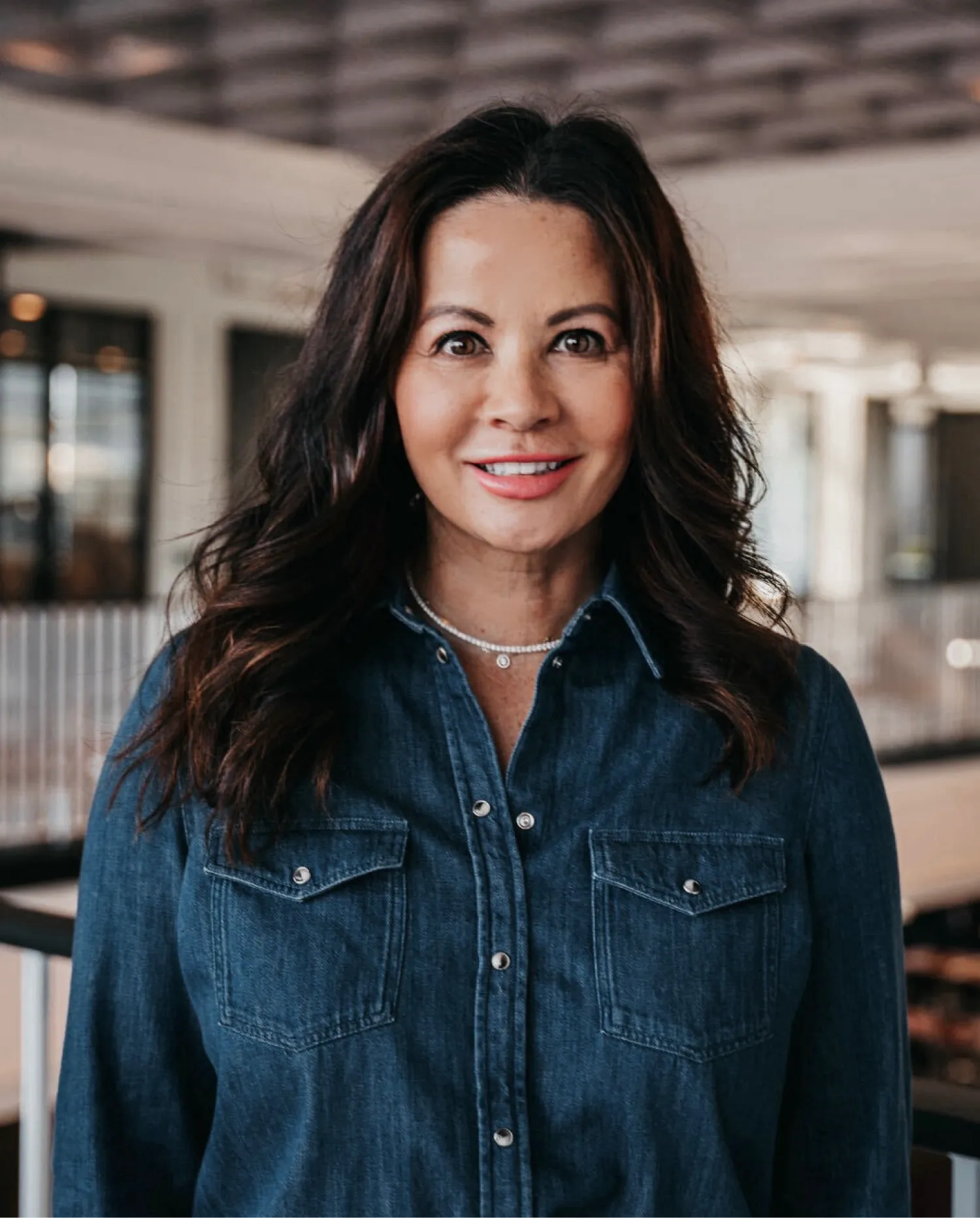 Smiling woman with long brown hair wearing a dark denim button-up shirt in a modern office buillding.