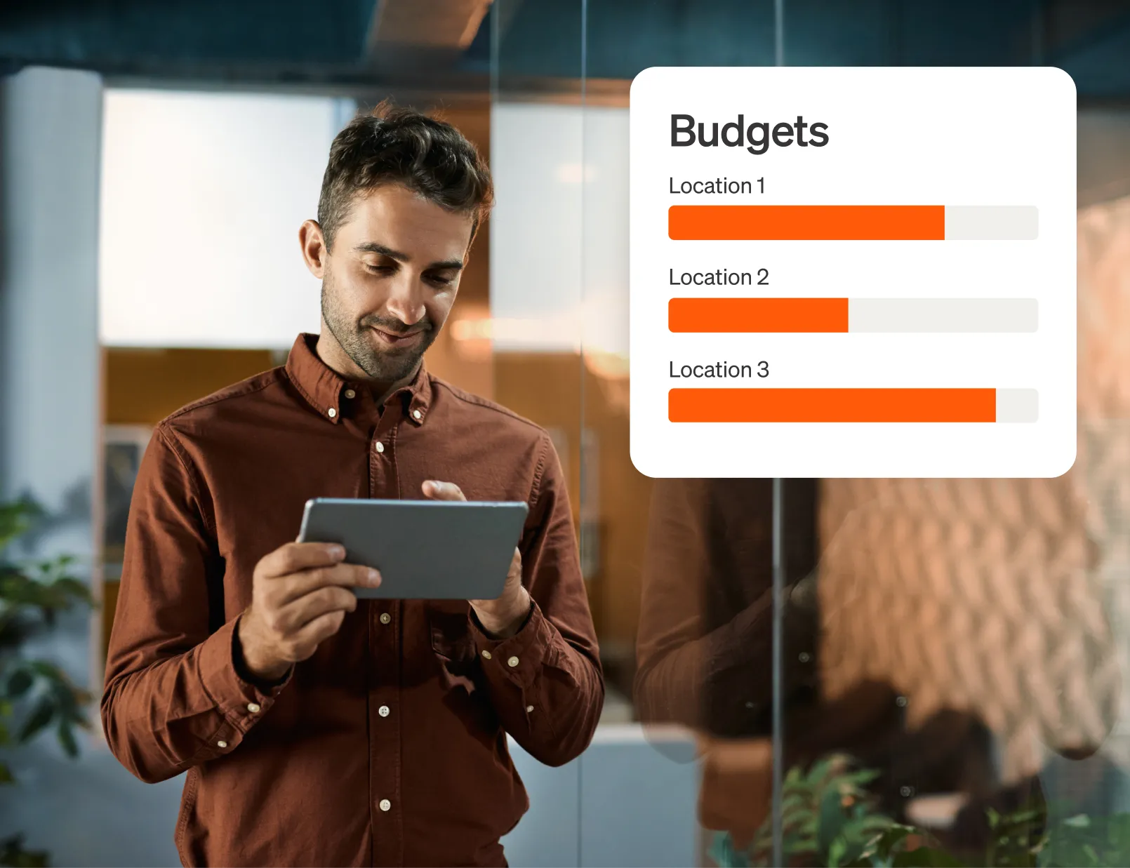 Man in brown button-down shirt stands in an office with a tablet in his hand, a UI screen for Budgets showing three locations with status bars for each.