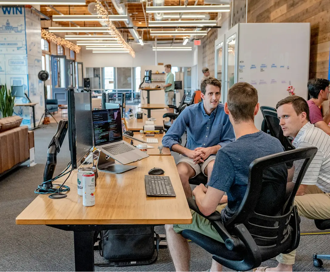 Three men seated and talking at a wooden desk with computer monitors and a laptop in a modern office space.