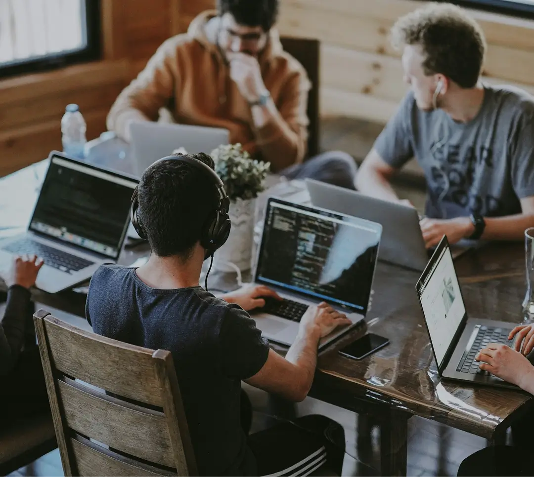 Group of young people working on laptops around a wooden table in a cozy room.