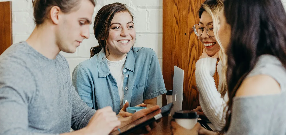 Four young adults sitting around a table in a casual setting, engaged in conversation and working on digital devices.