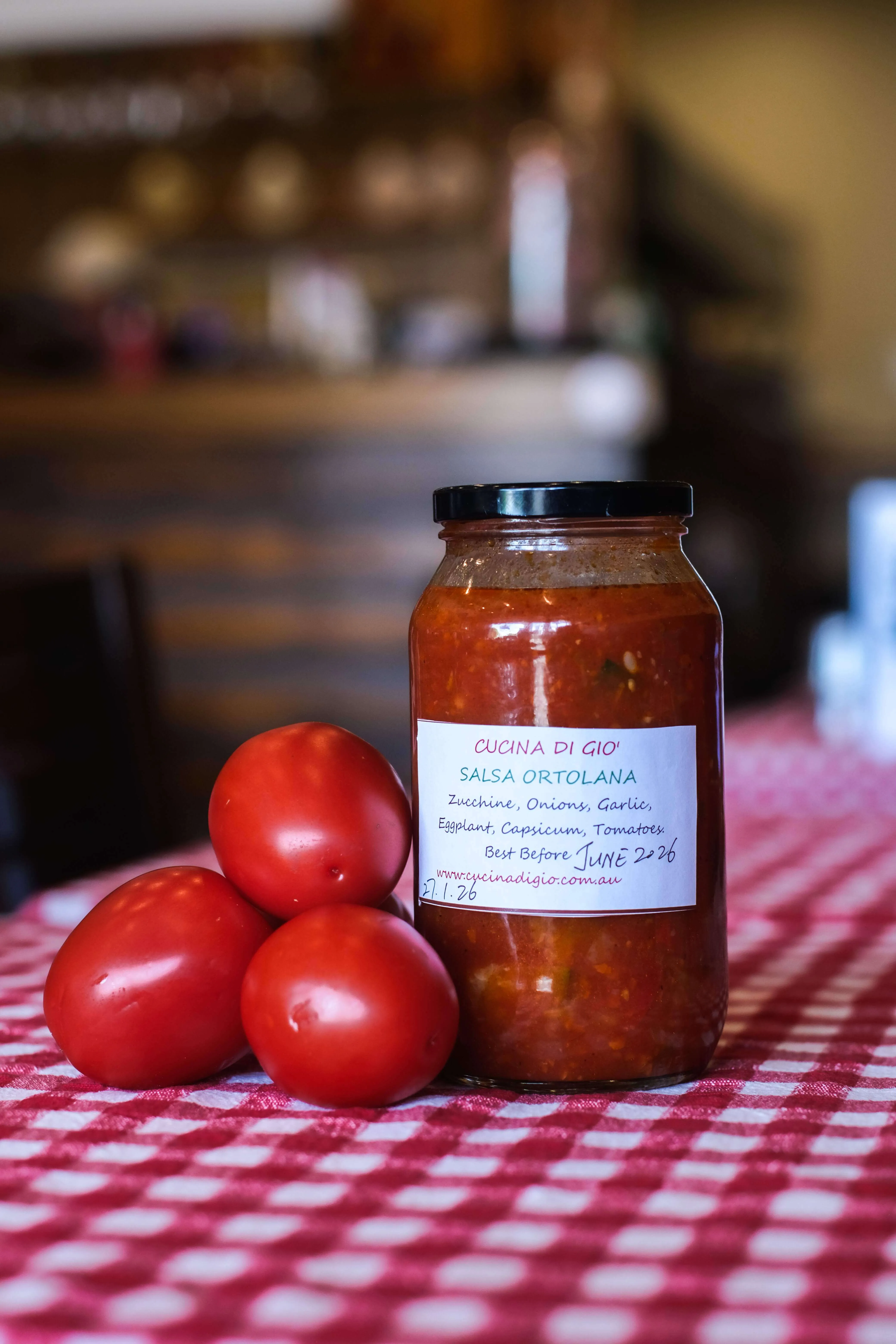 Jar of beef ragu sauce with three fresh tomatoes on a red and white checkered tablecloth, with a Cocina Di Gio wine list card in the background.