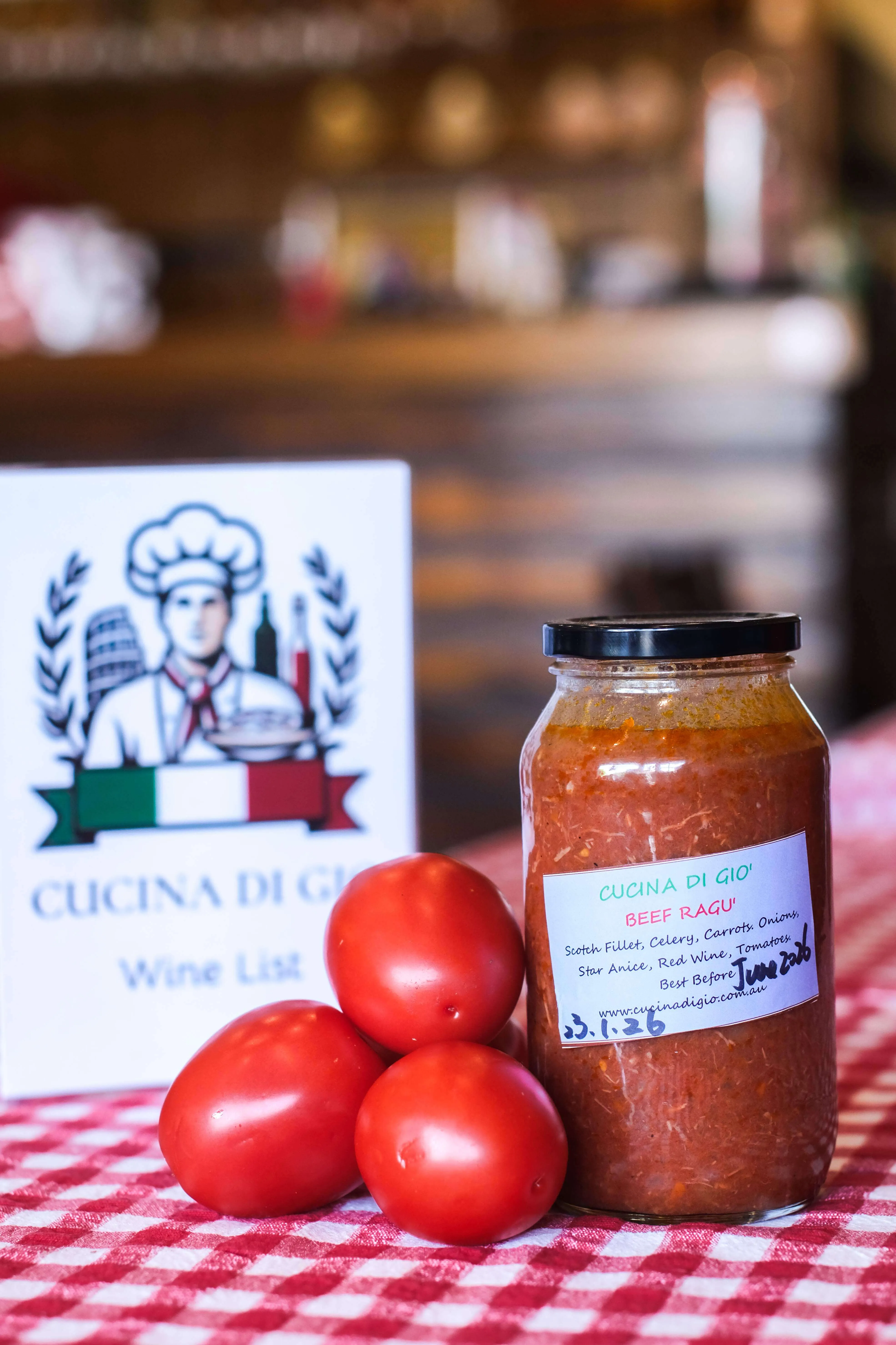 Jar of beef ragu sauce with three fresh tomatoes on a red and white checkered tablecloth, with a Cocina Di Gio wine list card in the background.