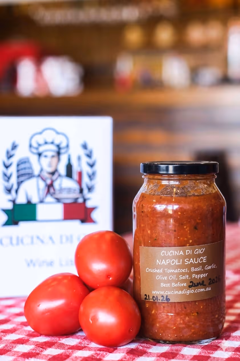Jar of beef ragu sauce with three fresh tomatoes on a red and white checkered tablecloth, with a Cocina Di Gio wine list card in the background.