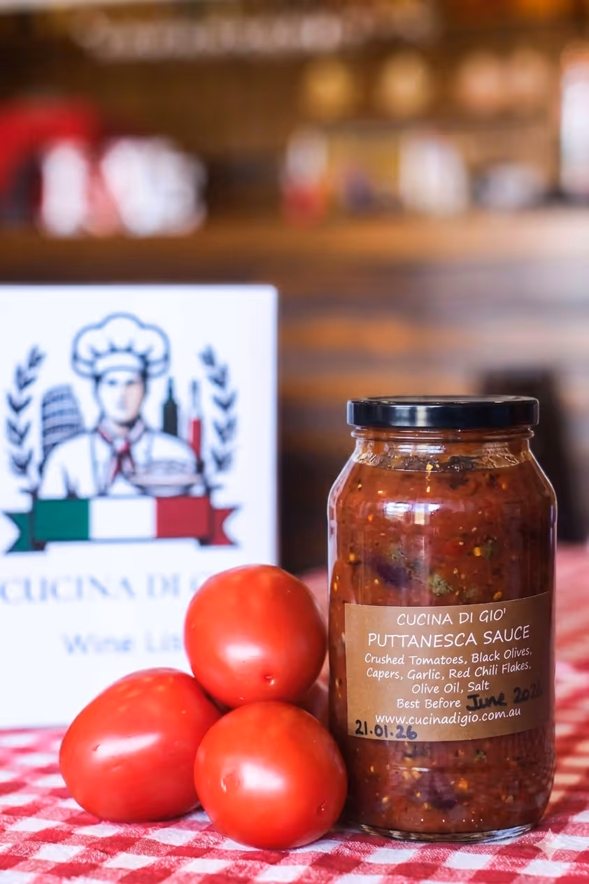 Jar of beef ragu sauce with three fresh tomatoes on a red and white checkered tablecloth, with a Cocina Di Gio wine list card in the background.