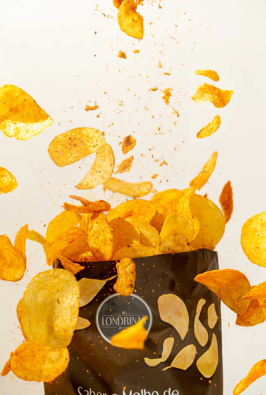 Pacote de batatas fritas sabor a molho de francesinha da marca Londrina Chips douradas temperadas caindo e saindo de um pacote preto de batatas fritas Londrina's.