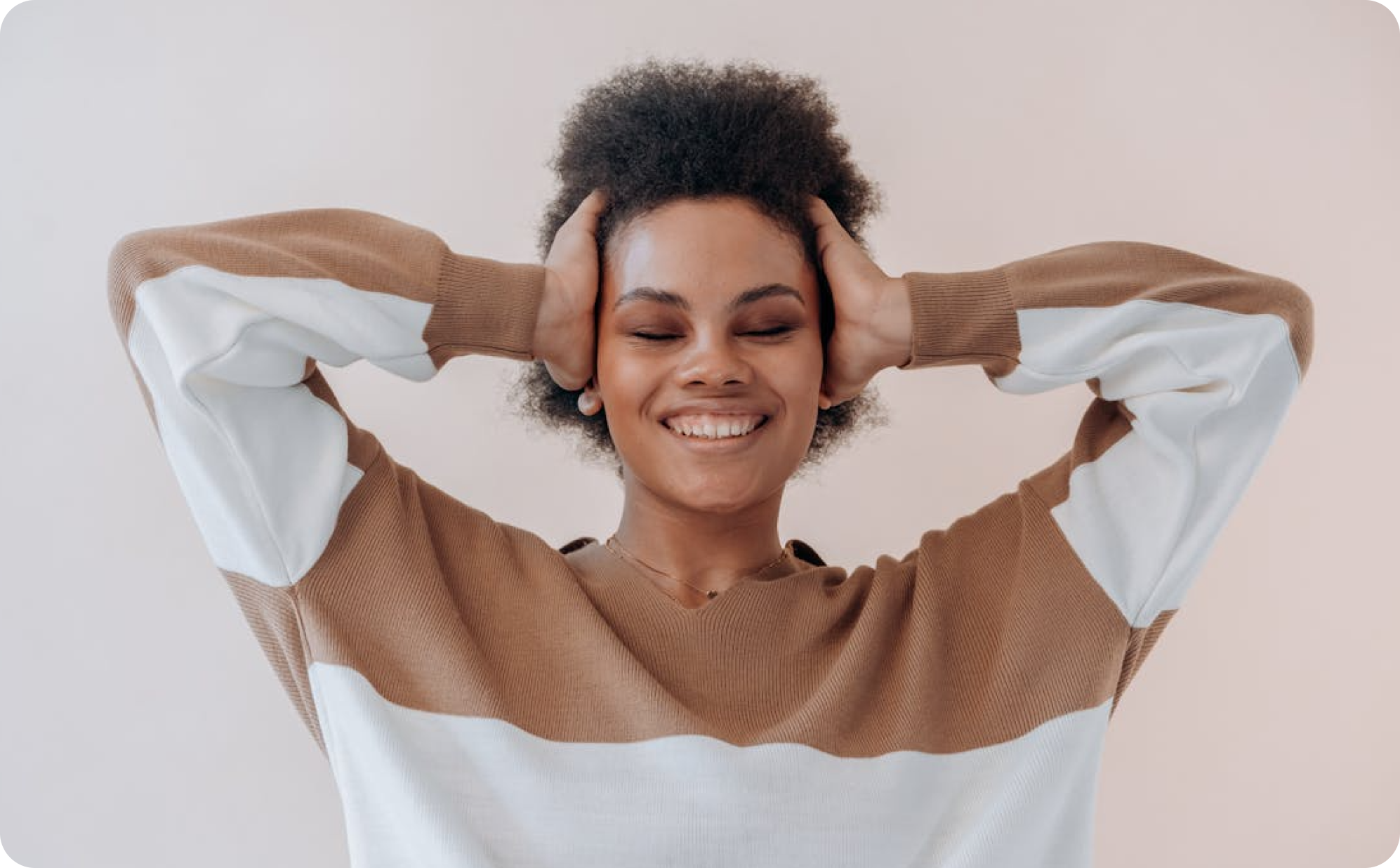 African American woman smiling with eyes closed and hands pressed against her head, beige background.