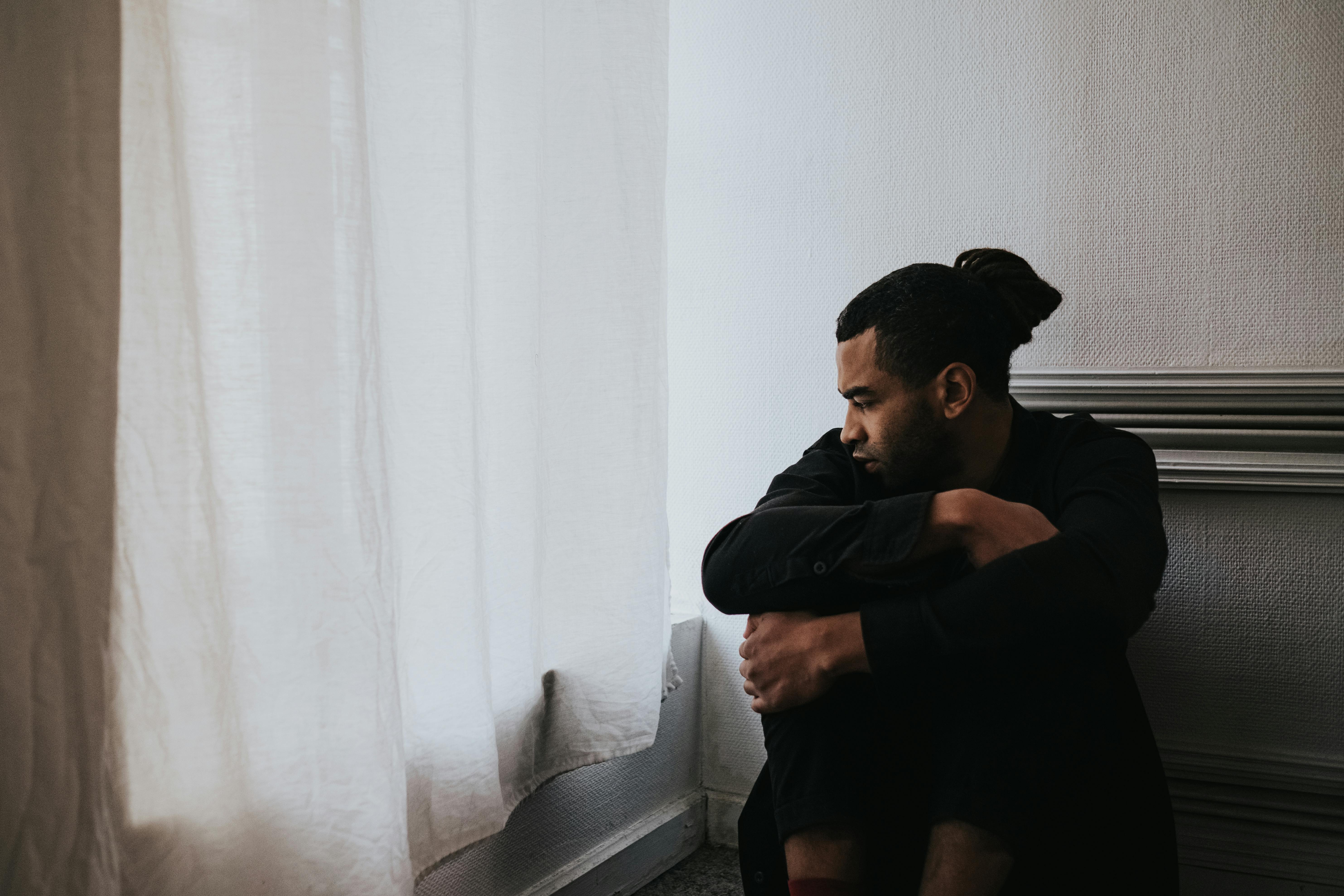 Black man sitting by a window covered by a sheer white curtain, looking sad