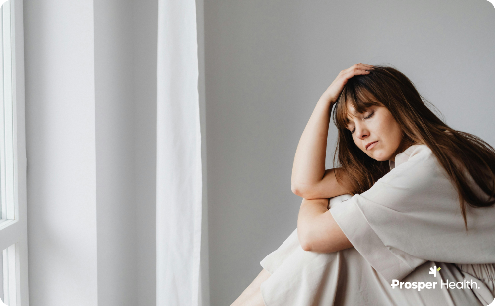 white woman with long brown here woman sitting in a white room with her hand on her head and her eyes closed