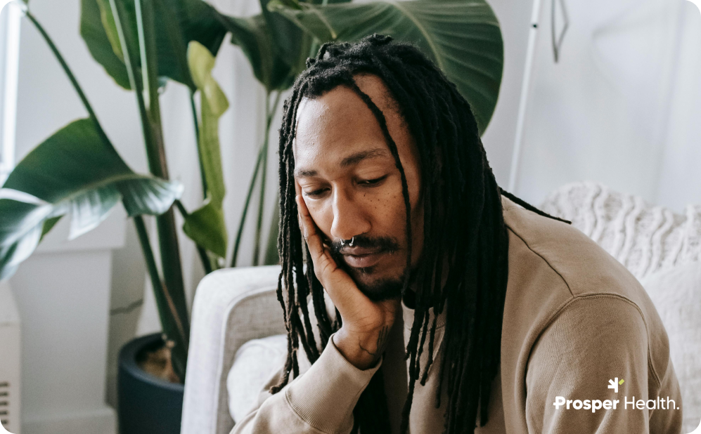 Black man with dreadlocks sitting in a room with a plant placing his head in his hand