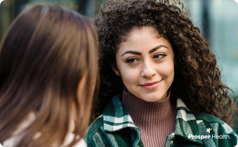 Woman with black curly hair looking at her friend, who has her back to the camera.