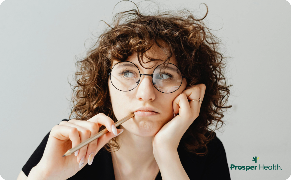 young woman with curly hair and round glasses thinking, putting her pencil up to her mouth