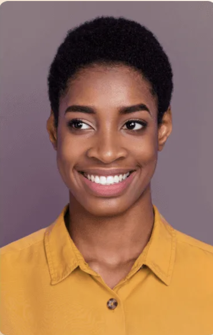 Smiling woman with short curly hair wearing a mustard yellow collared shirt against a muted purple background.