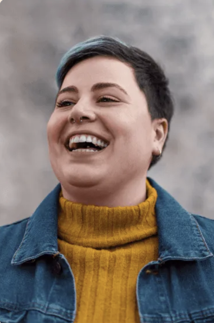 Person with short dark hair and a blue streak, wearing a mustard yellow turtleneck and blue denim jacket, smiling and looking slightly to the side.