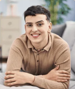 Young person with short dark hair wearing a light brown ribbed sweater, smiling and leaning forward with arms crossed.