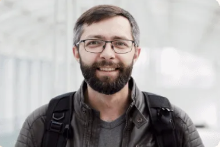Smiling man with glasses and a beard wearing a gray jacket and backpack.