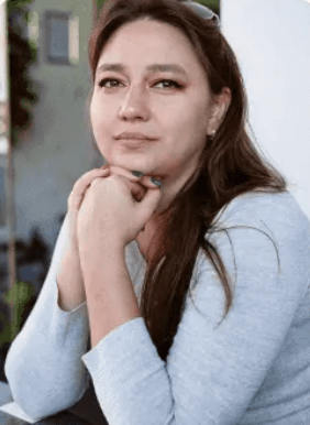 Woman with long brown hair resting her chin on her clasped hands, wearing a light gray sweater and sitting indoors.