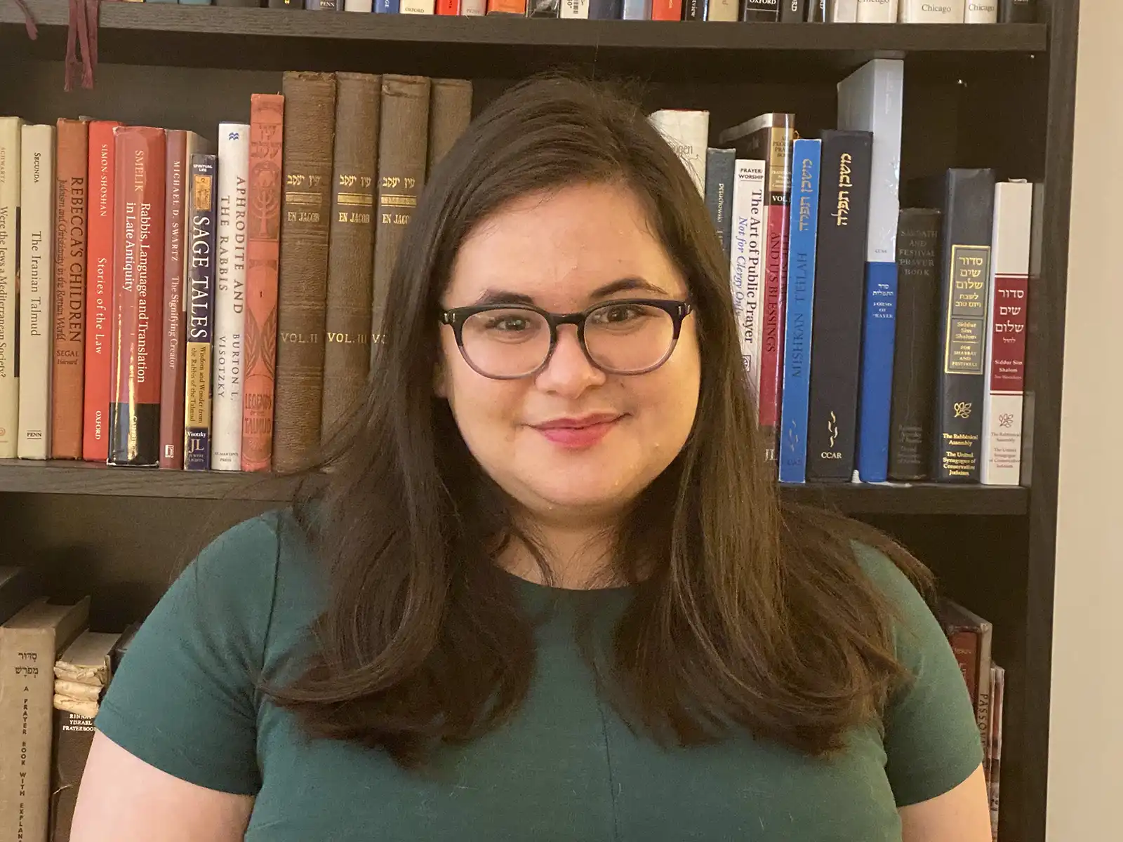 Annie Norman-Schiff with long dark hair in a green shirt standing in front of a bookshelf filled with various books in a home or office setting.