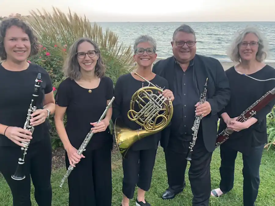 The Elm City Winds pose outdoors by the water holding woodwind and brass instruments, including clarinet, flute, French horn, oboe, and bassoon.