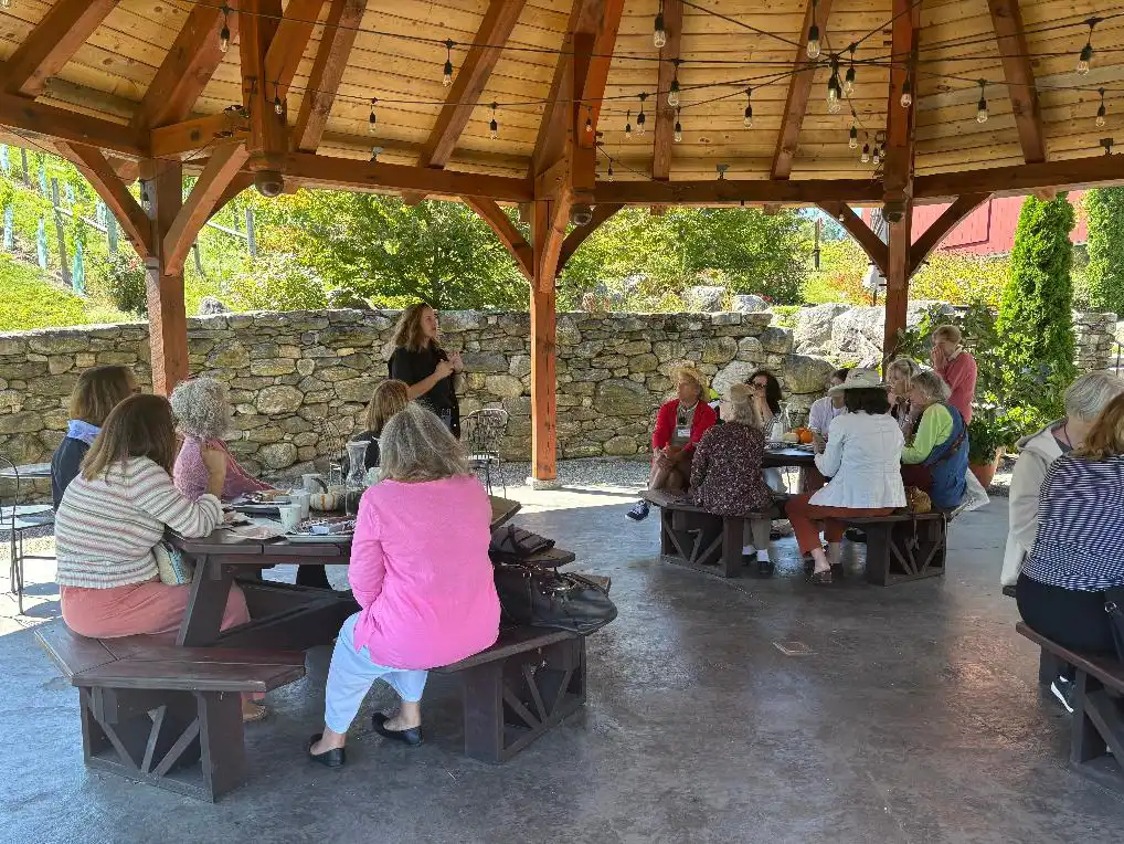 Garden Club members enjoying a charcuterie lunch under a large wooden gazebo at Jones Vineyards, with the owner giving a winery presentation.