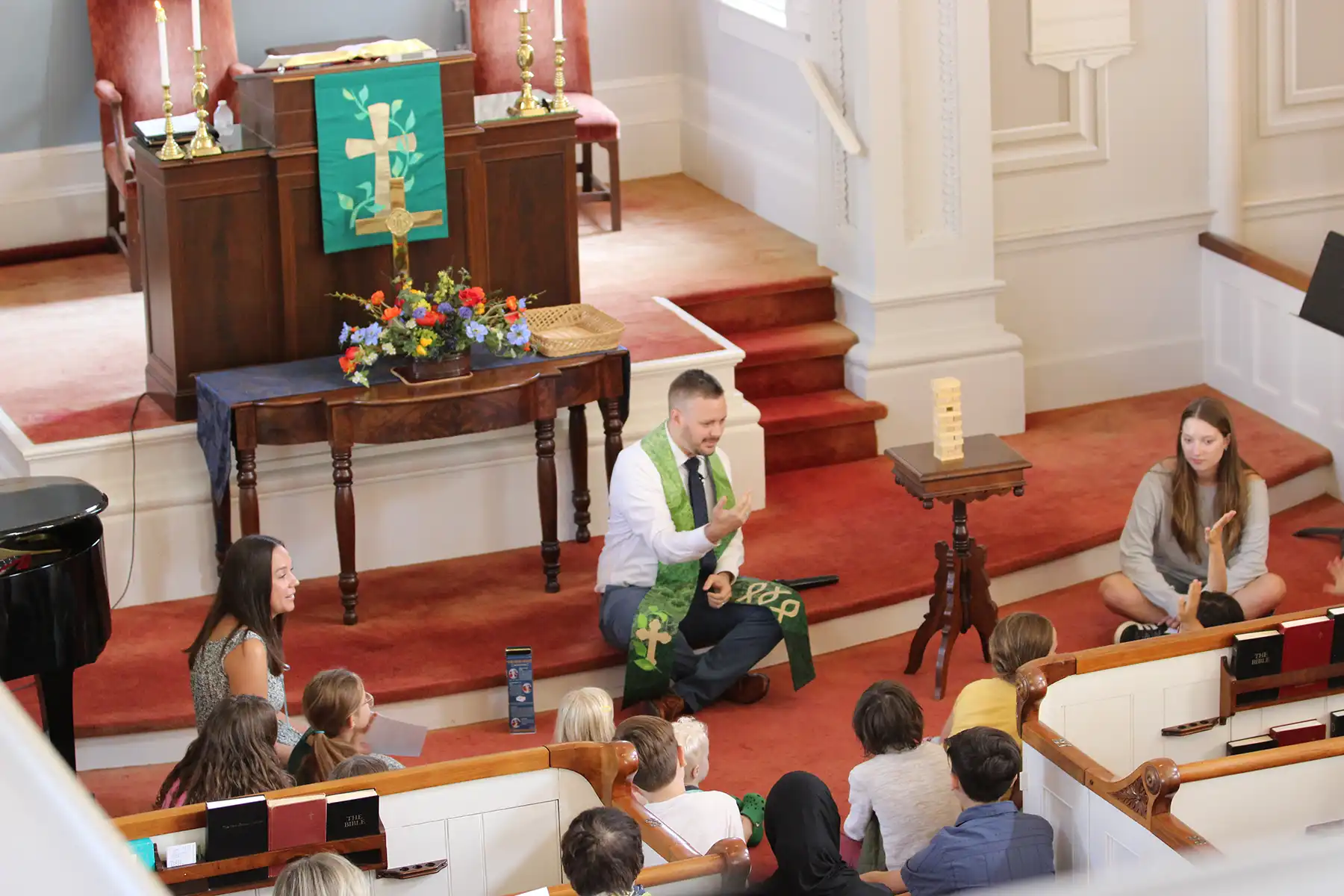 Pastor Andrew sits on the church floor, teaching a group of children during a Sunday morning service, with adults and kids listening attentively around him.