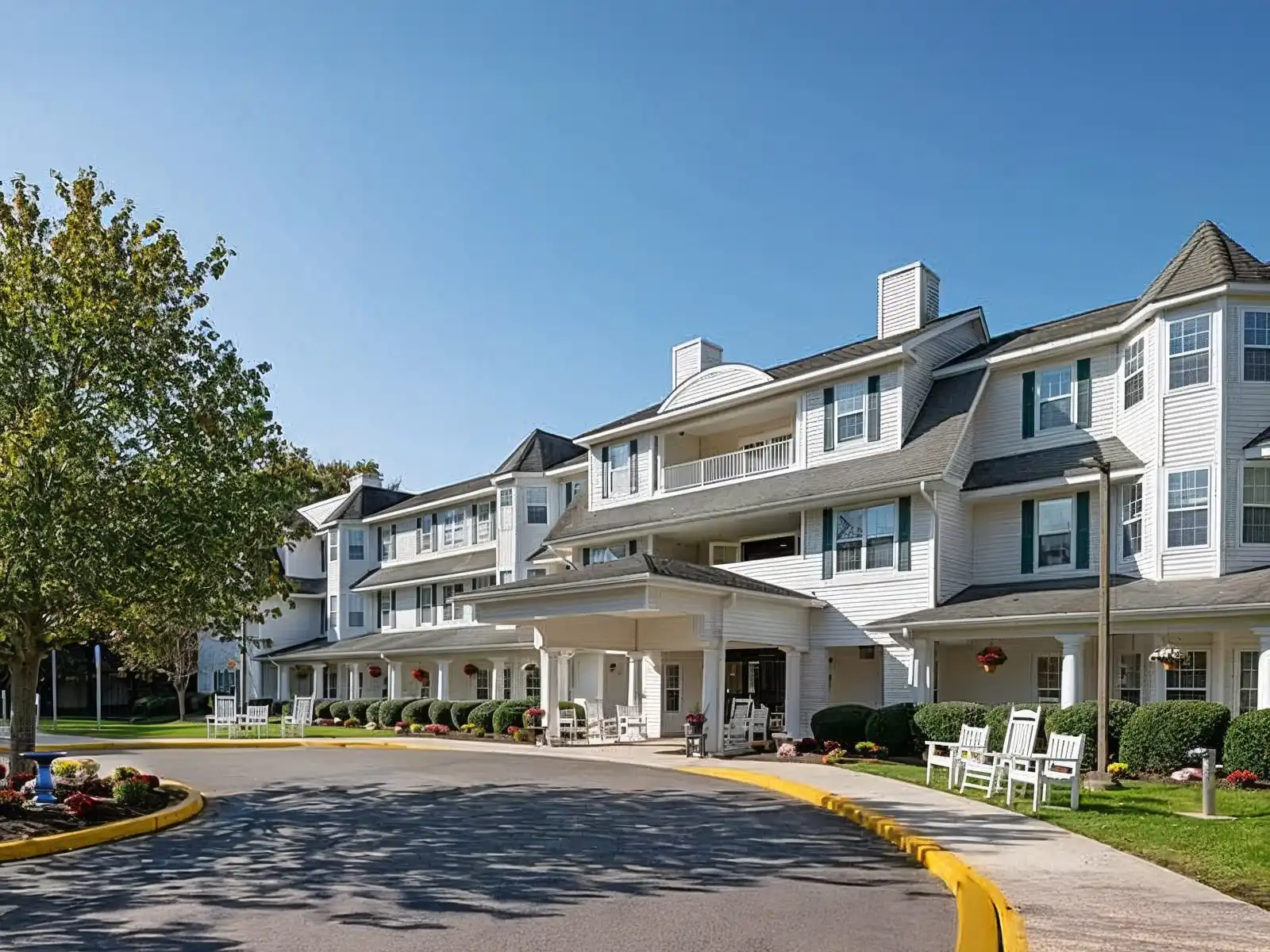 Front view of Charter of Woodbridge senior living community showing white building exterior, landscaped gardens, benches, and clear blue sky.