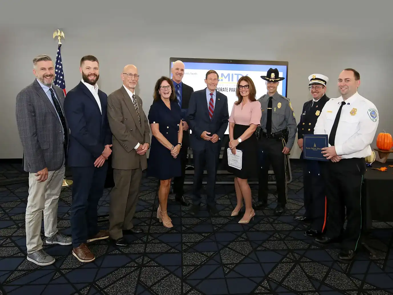 A group photo of Amity Chamber Community Awards winners, police, firefighters, and officials standing indoors in front of a display and American flag.