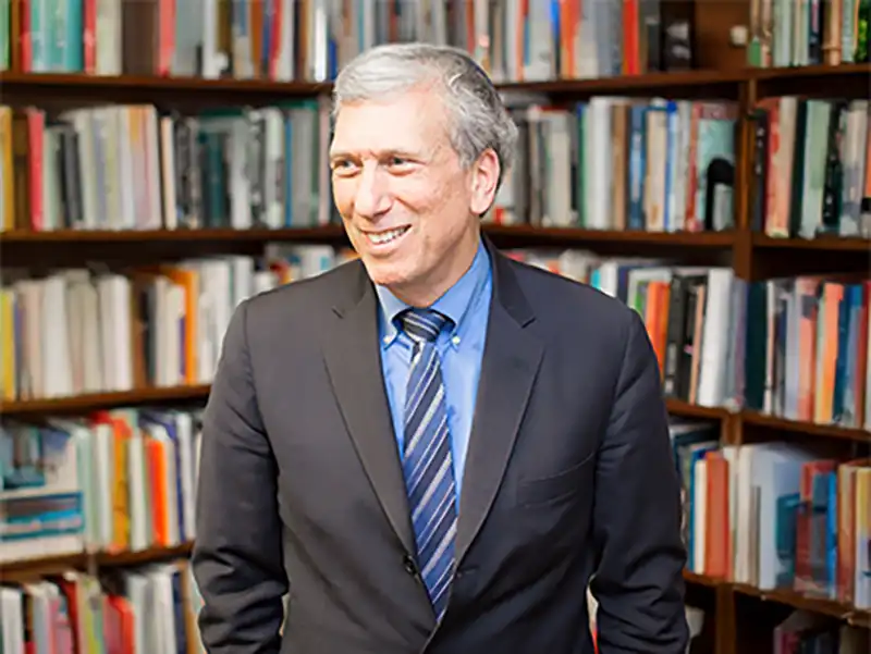 Professional headshot of Professor Arnold Eisen in a dark suit, light blue shirt, and striped tie, standing in front of a full bookshelf.