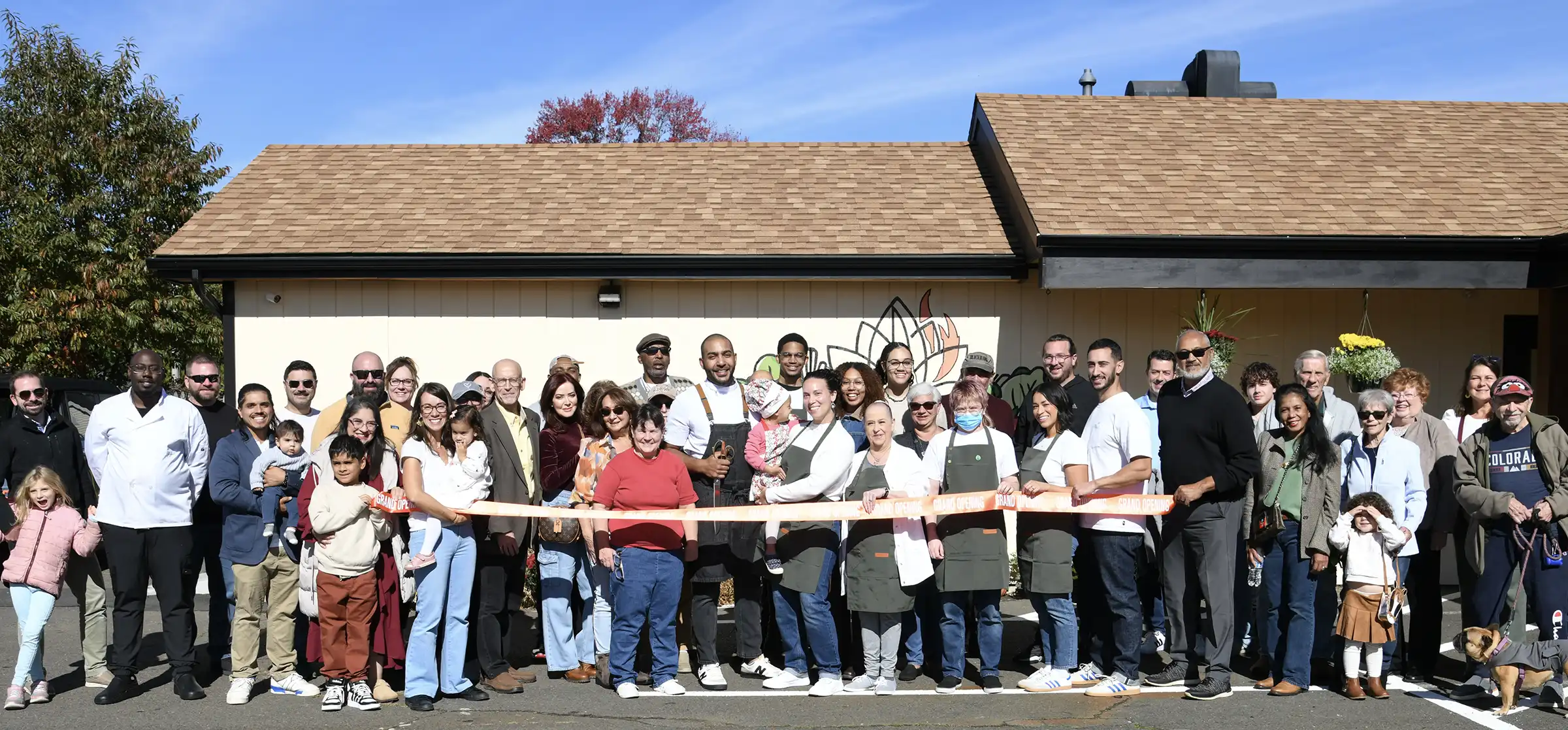A diverse group of over 30 adults and children, including staff in aprons, pose smiling outdoors holding a ribbon for a Mantras Market grand opening.