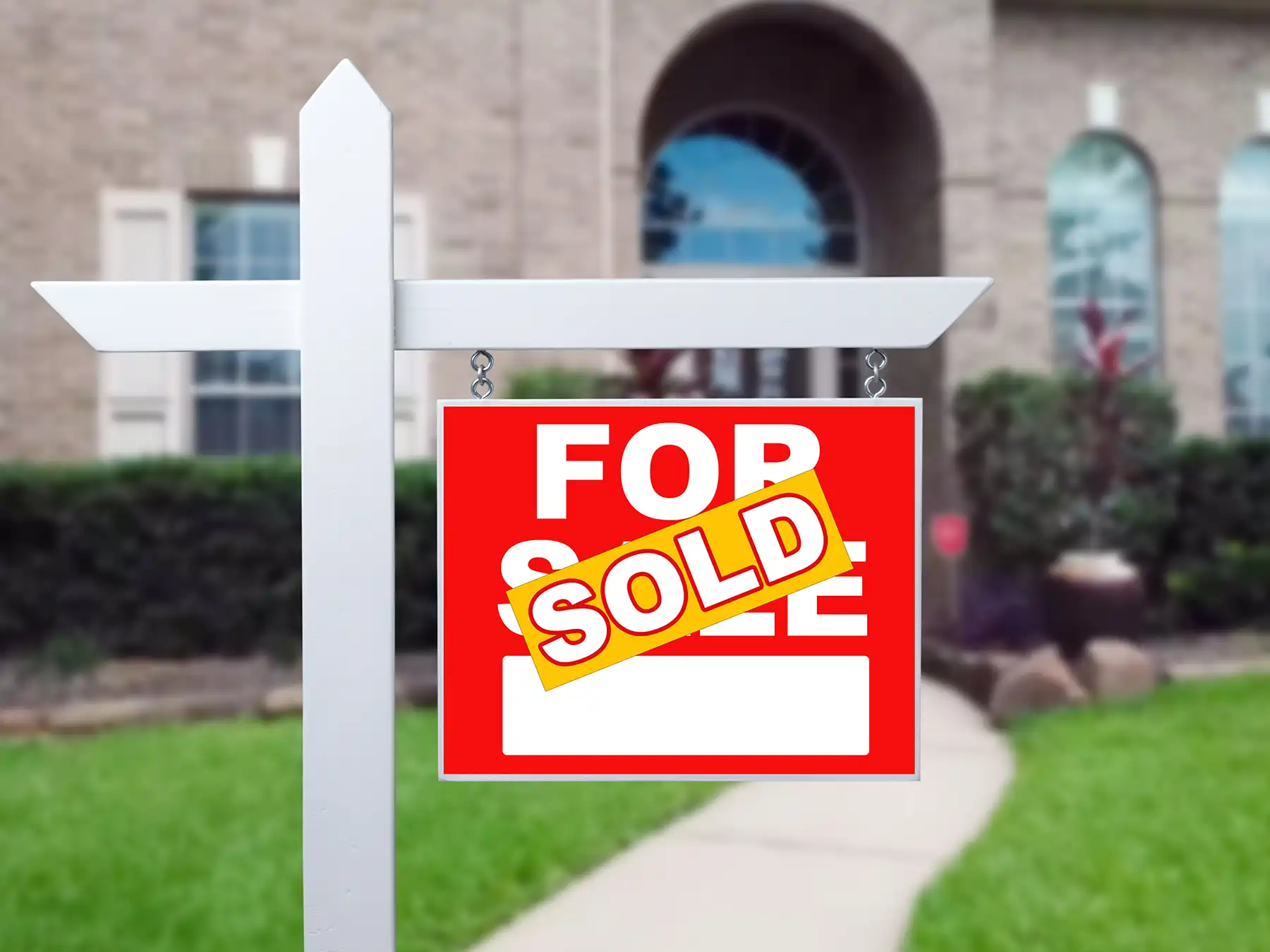 Home 'For Sale' sign with a yellow 'SOLD' banner over the red card, planted in the lawn near a walkway to a blurred brick house.