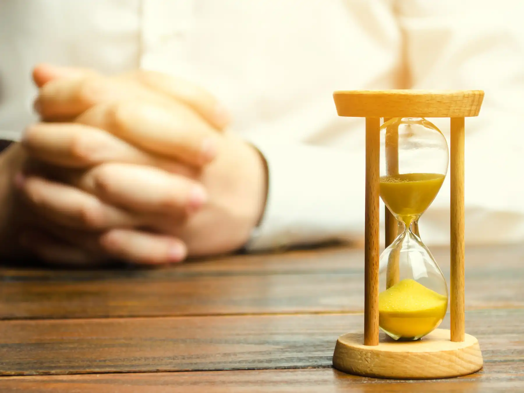 A person sits with hands folded on a wooden table, focusing on a yellow sand hourglass, symbolizing waiting, passing time, or decision-making.
