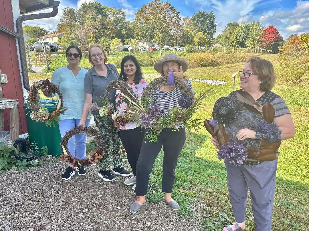 Five Garden Club members display handmade grapevine wreaths outdoors at Massaro Farm, decorated with hydrangeas, pine cones, and various natural florals.