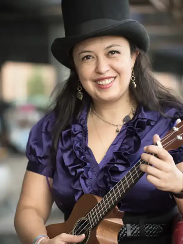 Smiling woman, Rachel Haymer, in a top hat and purple ruffled blouse, playing a ukulele, possibly a musician or entertainer.