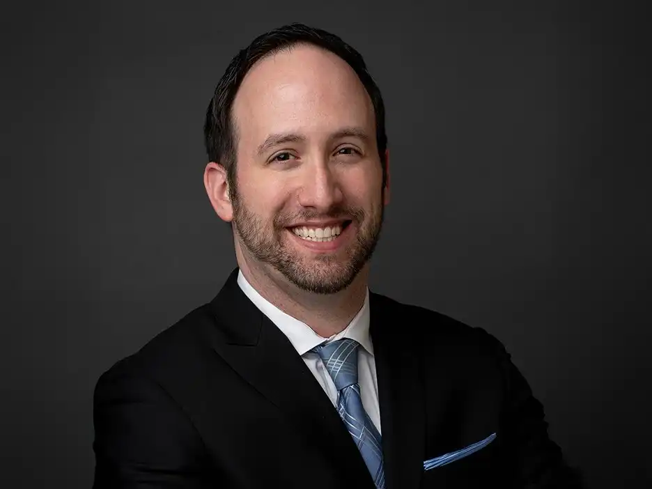 Close-up professional headshot of Owen Weaver smiling, wearing a dark suit with a light blue patterned tie and pocket square against a gray backdrop.
