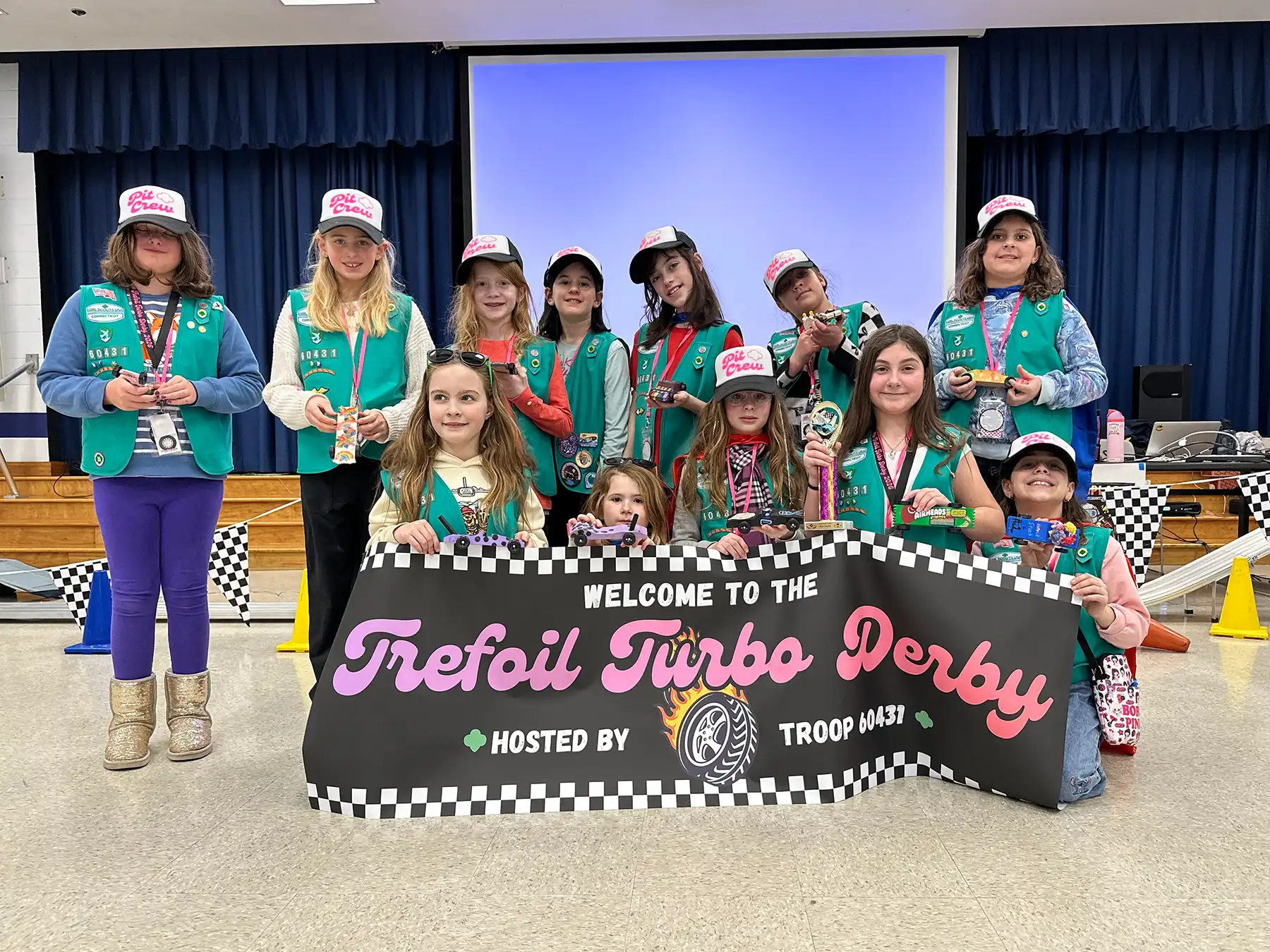 Girl Scout Troop 60431 members pose with pinewood derby cars and a "Trefoil Turbo Derby" banner at a racing event, wearing teal vests and pink pit crew hats.