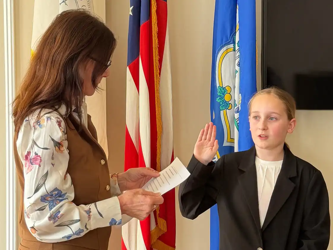 A woman administers an oath of office to a young girl in a suit jacket raising her hand. US and Connecticut state flags are in the background.