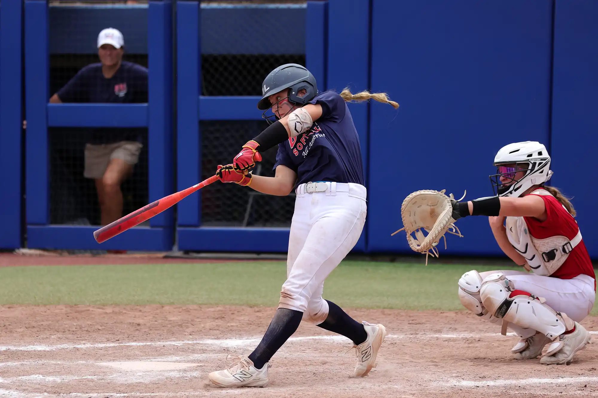 A softball player in a navy blue jersey hits a ball with a red bat while the catcher crouches behind her and an umpire watches from the dugout in the background