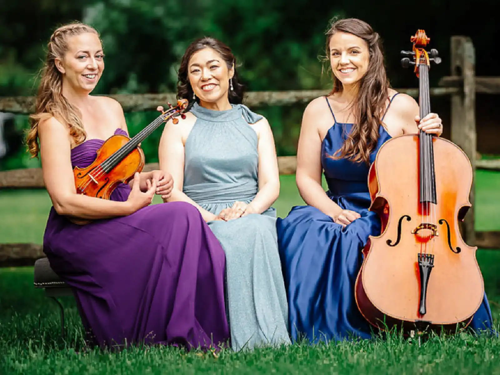 Three women of the Champlain Trio sit outdoors on a bench with a violin, piano, and cello, wearing elegant purple, grey, and blue dresses.