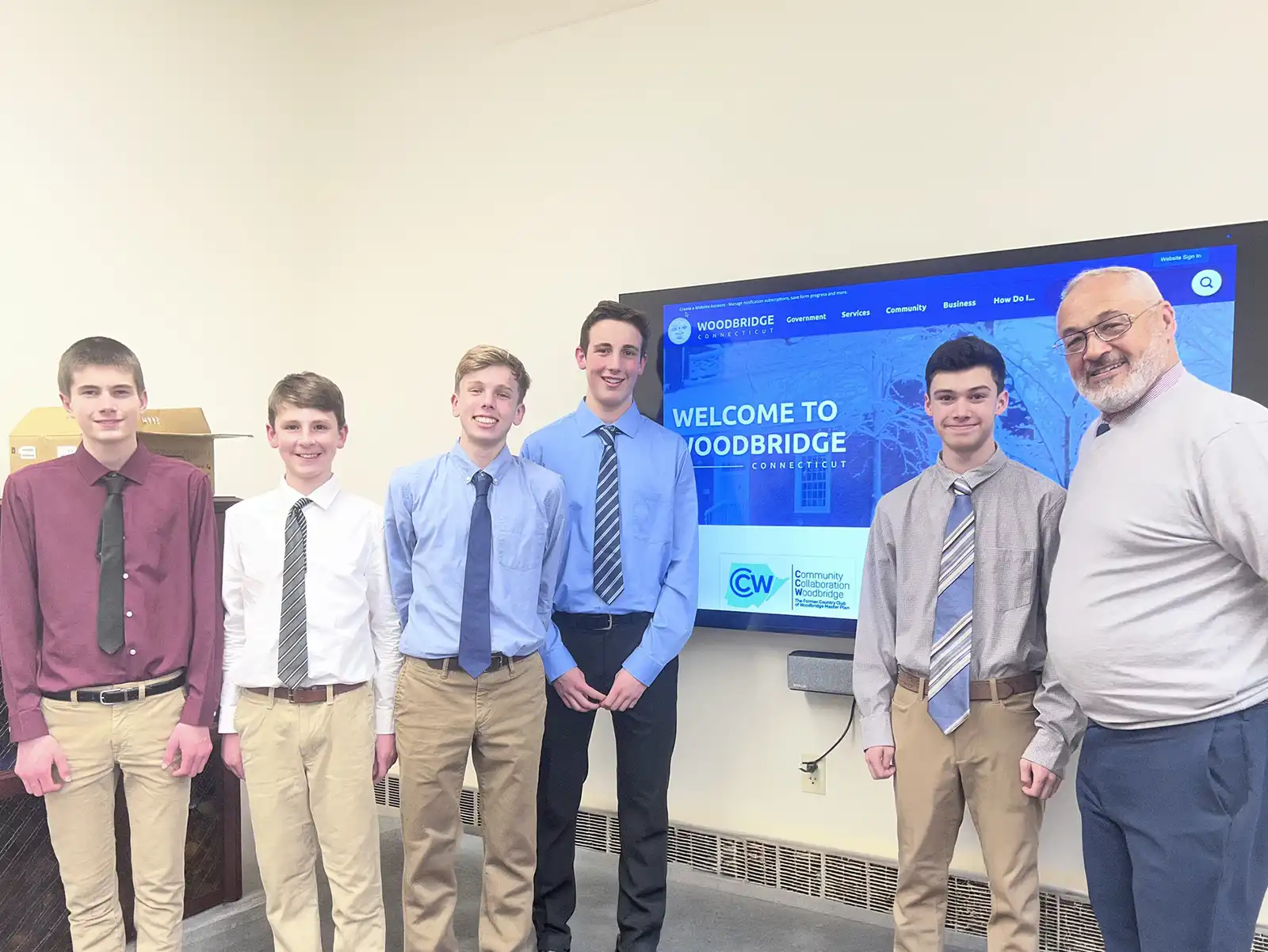 Six individuals pose for a group photo, with five students in shirts and ties and one adult, by a "Welcome to Woodbridge" screen.