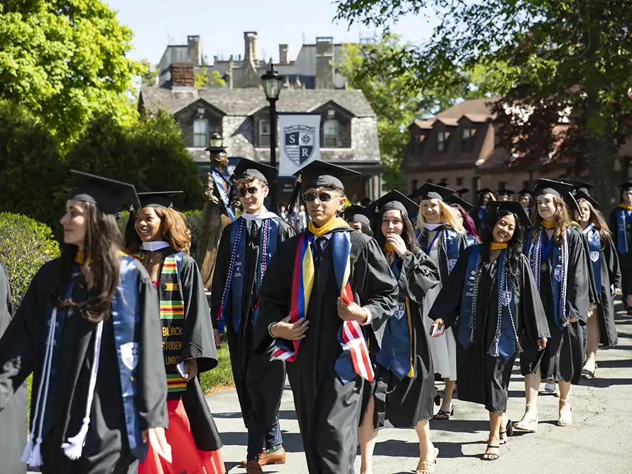 Graduates in black caps and gowns walk in an outdoor commencement procession on the Salve Regina University campus.