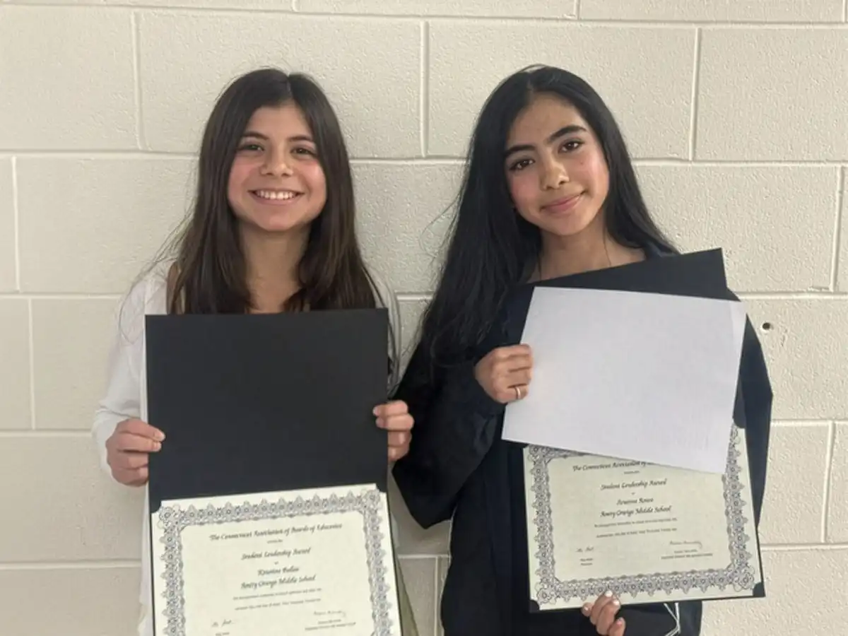 Two smiling 8th-grade girls stand in front of a white brick wall, proudly holding their CABE Student Leadership Award certificates.
