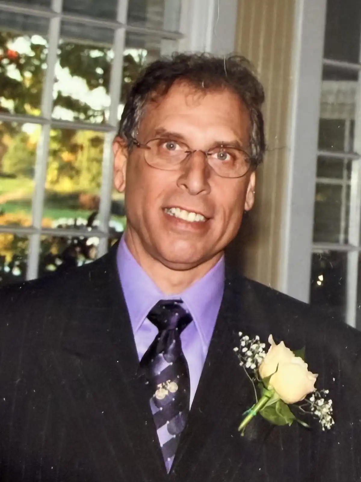 Portrait of Guy S. D’Aniello smiling in a black pinstripe suit, purple dress shirt, striped tie, and a rose boutonniere against a window.