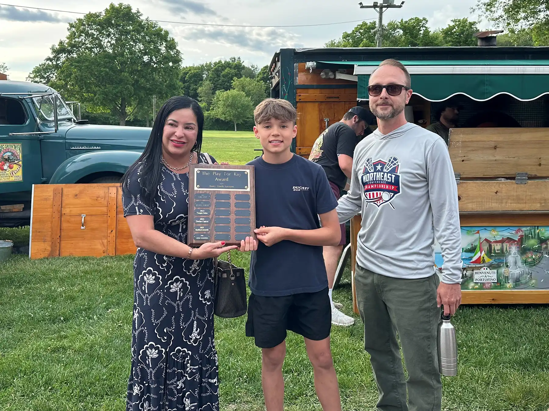 Marisol Heenie Sogluzzi, Lincoln Ruszkowski, and Eric Bowerman smile holding the Amity Youth Lacrosse Play For Ray Award plaque at an outdoor event.
