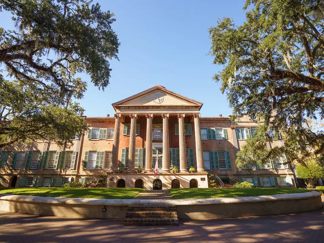 The historic College of Charleston Cistern Yard and Randolph Hall building featuring large columns, green shutters, and live oak trees.