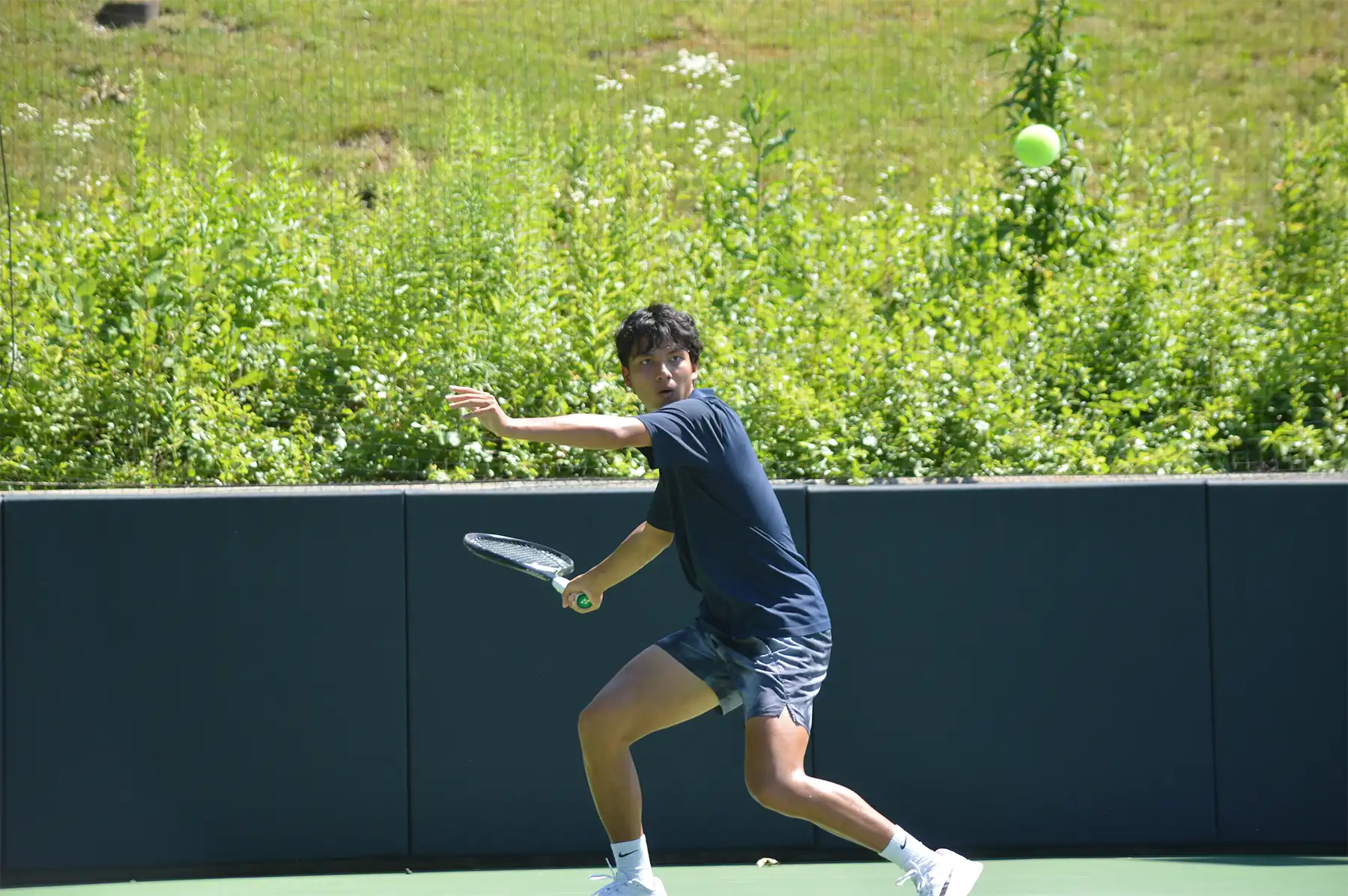 Amity High School tennis player Thomas Sirichantho tracking an incoming tennis ball to hit a backhand stroke on an outdoor court.