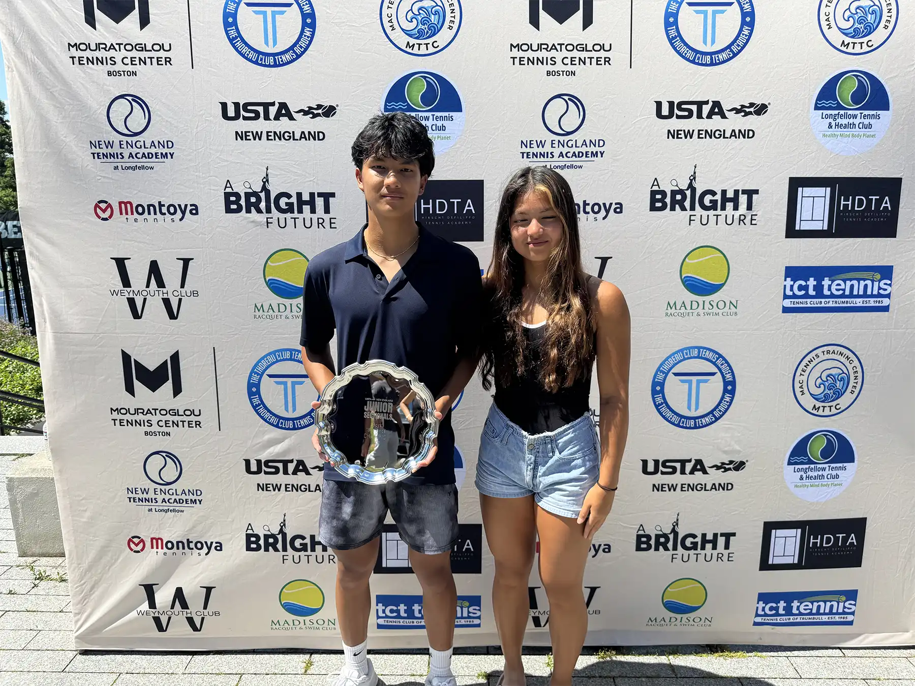 Thomas Sirichantho holds his New England Junior Tennis Sectionals trophy alongside his sister in front of a USTA backdrop.